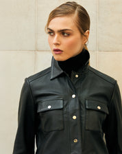 Close frontal da jaqueta de couro preta com gola clássica, bolsos de aba e botões de pressão metálicos, detalhe de costura aparente