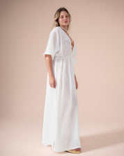 Vestido longo branco com mangas soltas e decote V, amarração abaixo do busto e fendas laterais, visto de perfil.