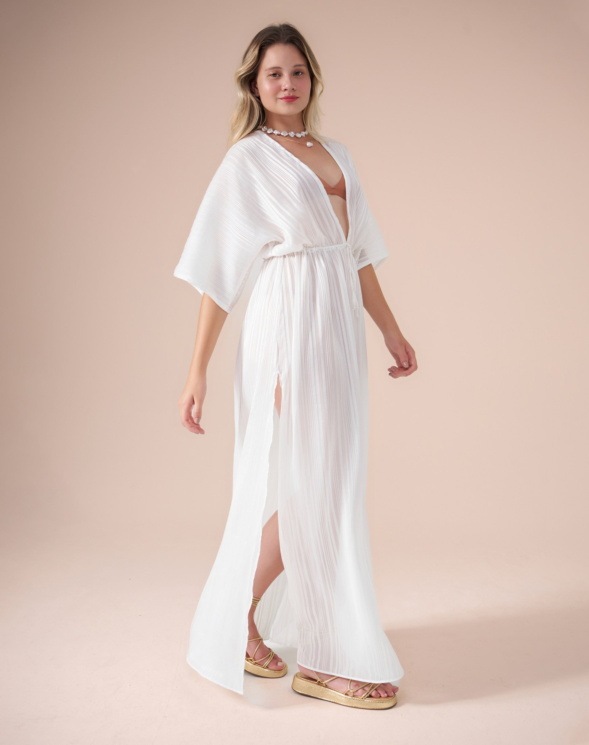 Vestido longo branco com mangas soltas e decote V, amarração abaixo do busto e fendas laterais, visto de perfil com movimento.