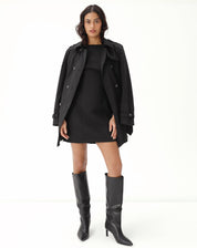 Modelo vestindo o vestido curto preto com botas e casaco, mostrando um look completo.