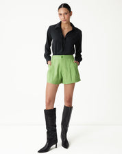 Modelo vestindo shorts verde com vincos e camisa preta, combinando com botas pretas.