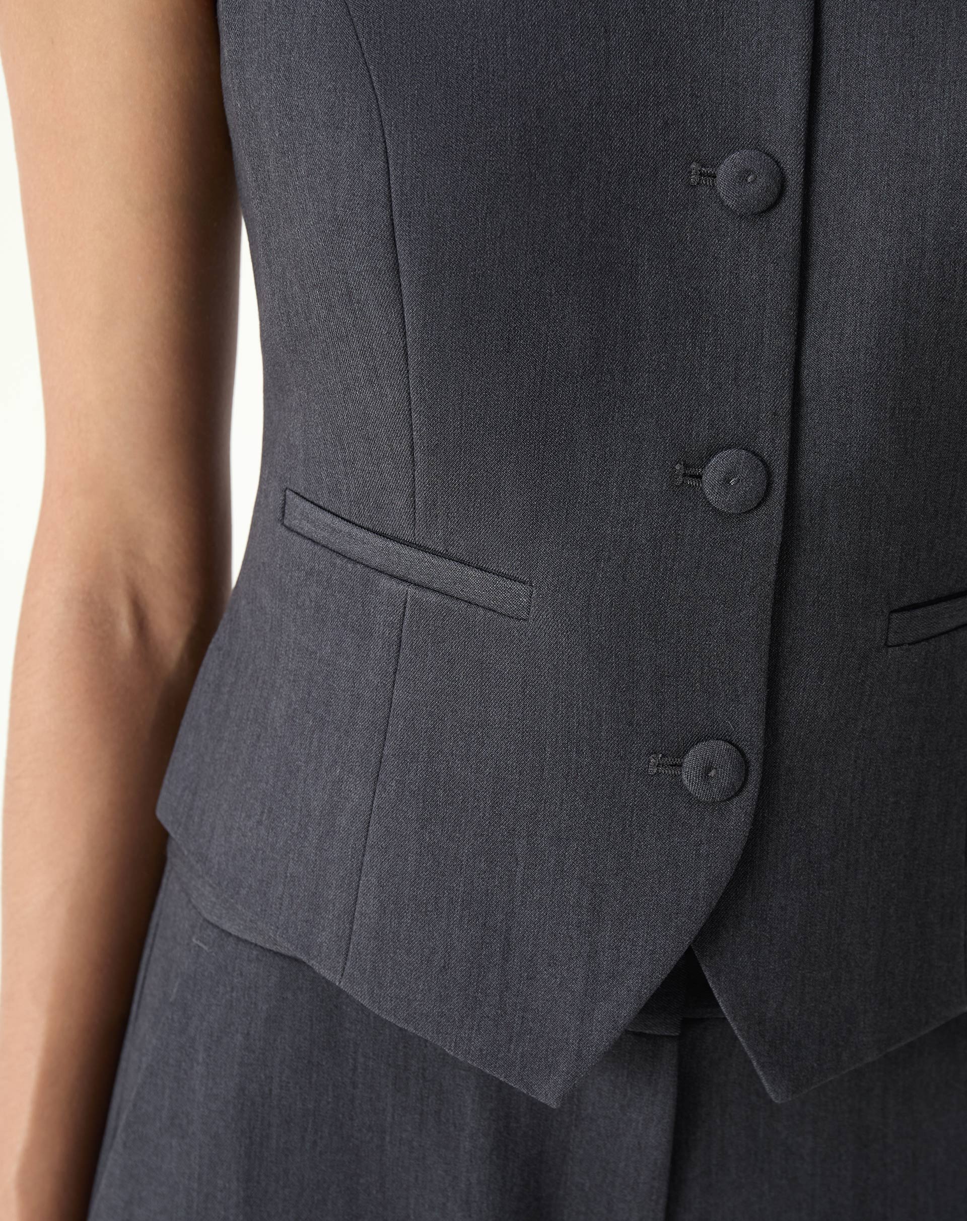 Detalhe do bolso embutido e dos botões forrados do colete alfaiataria cinza com acabamento visível.