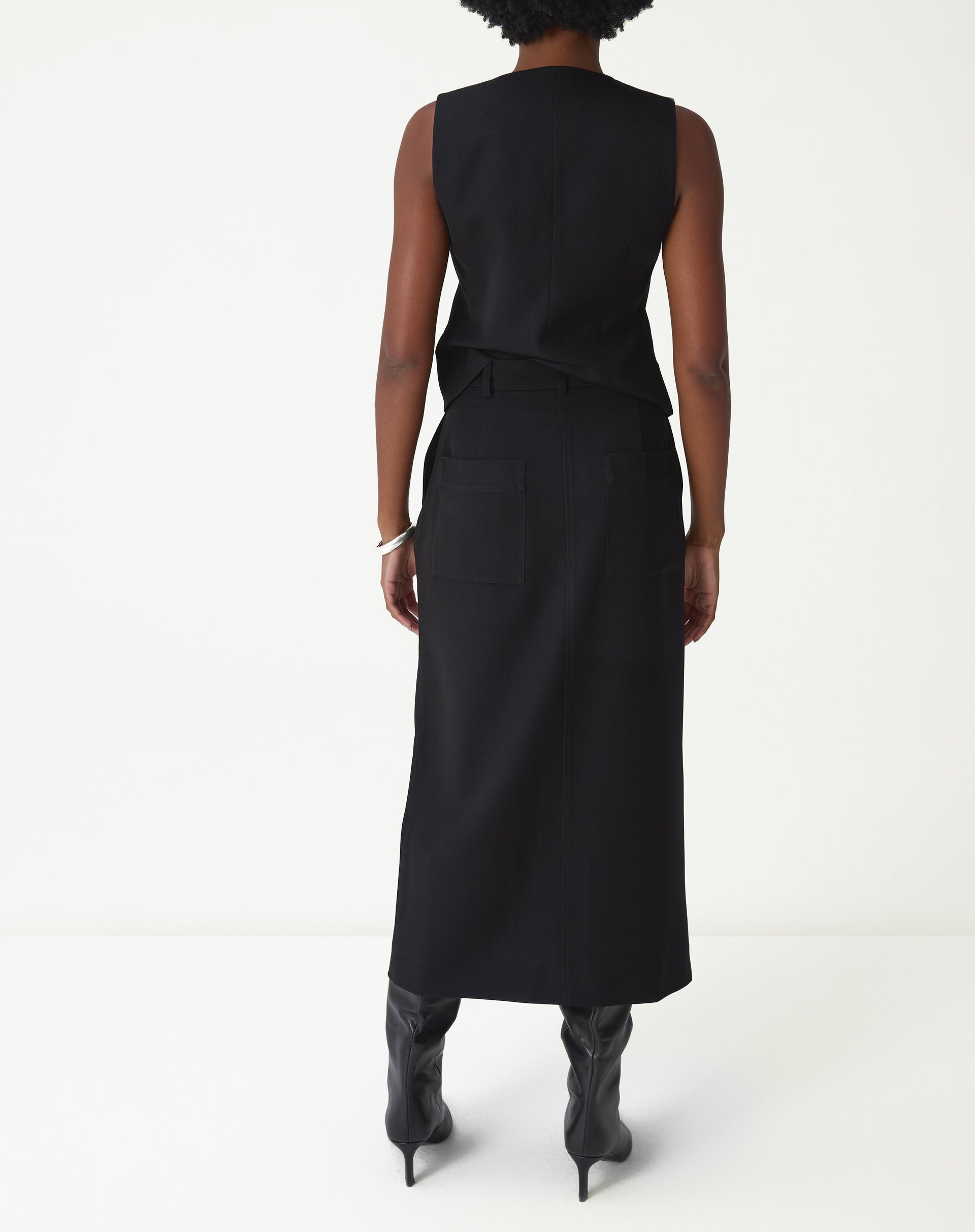 Vista traseira da modelo usando saia midi preta alfaiataria mostrando comprimento e caimento.
