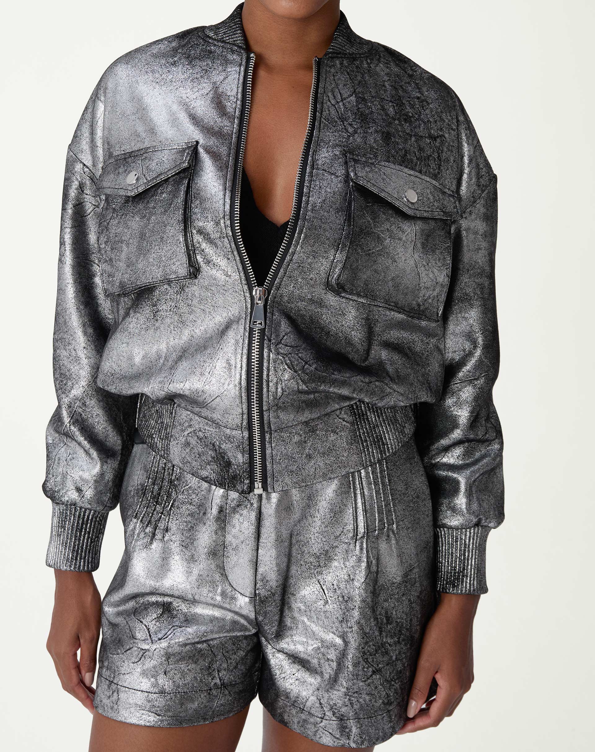 Modelo vestindo jaqueta bomber prata em suedine poliéster, vista frontal curta com zíper parcialmente aberto e shorts prateados combinando