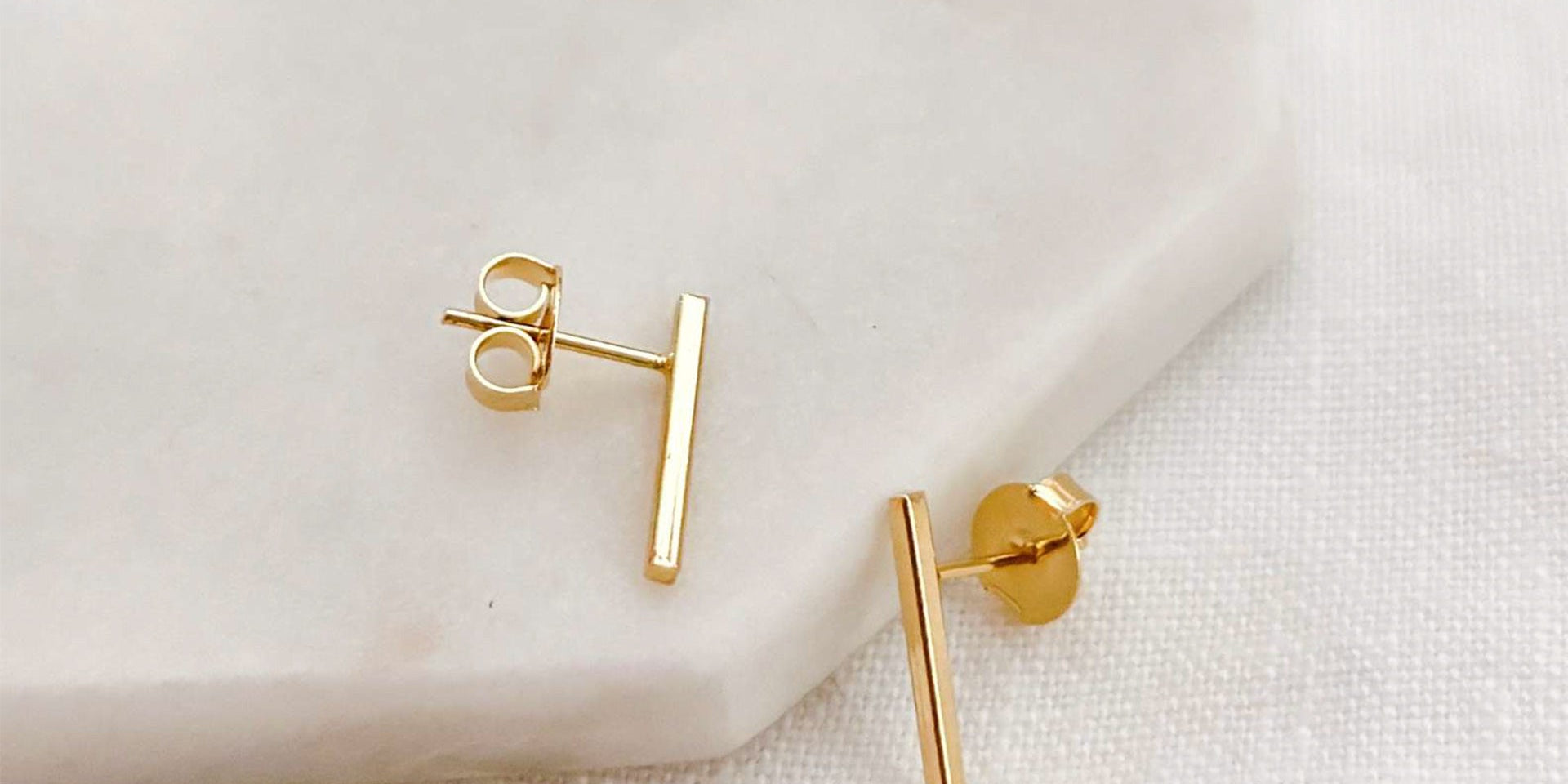 Brincos pequenos em formato de tubinho com acabamento liso e banho de ouro 18k sobre uma superfície branca.