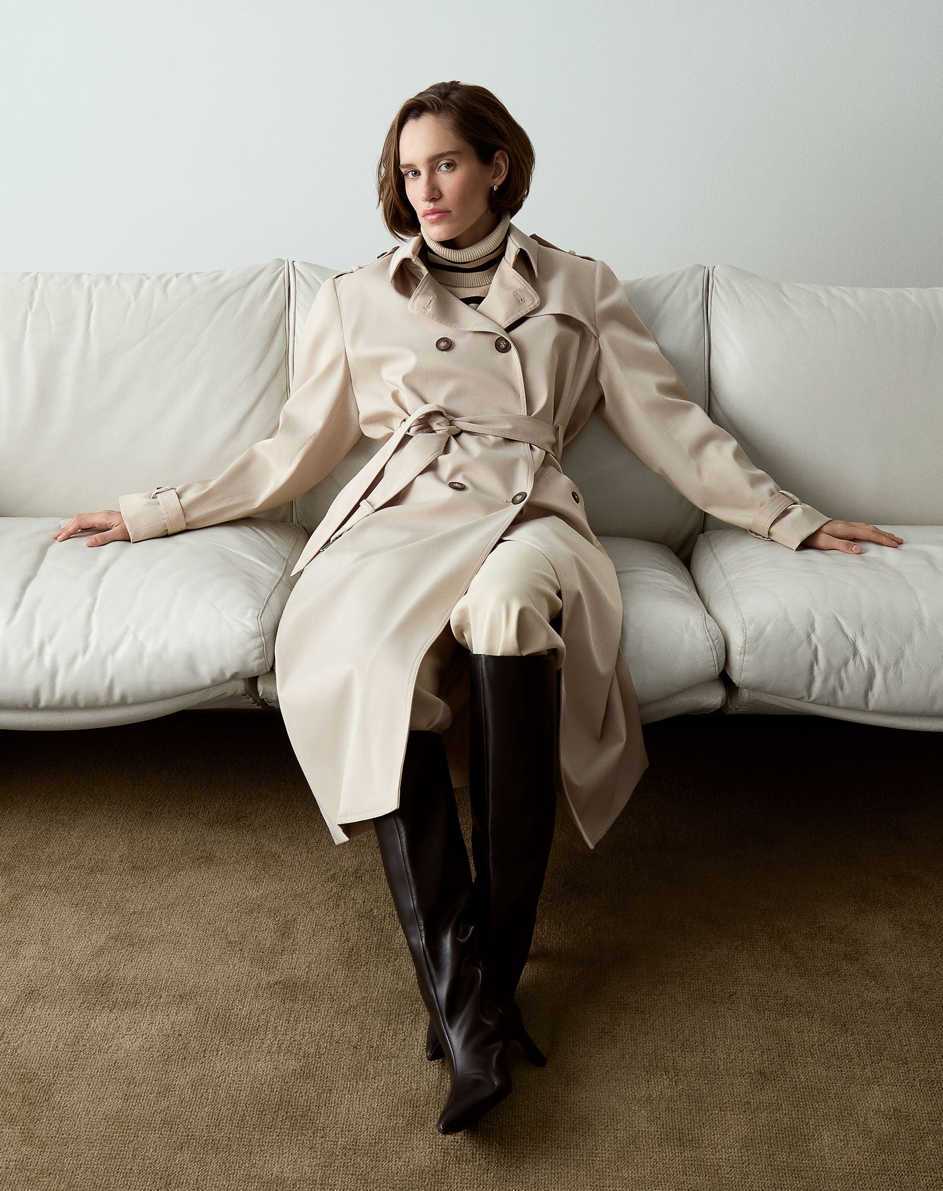 Modelo sentado em sofá vestindo trench coat bege com botas de couro.