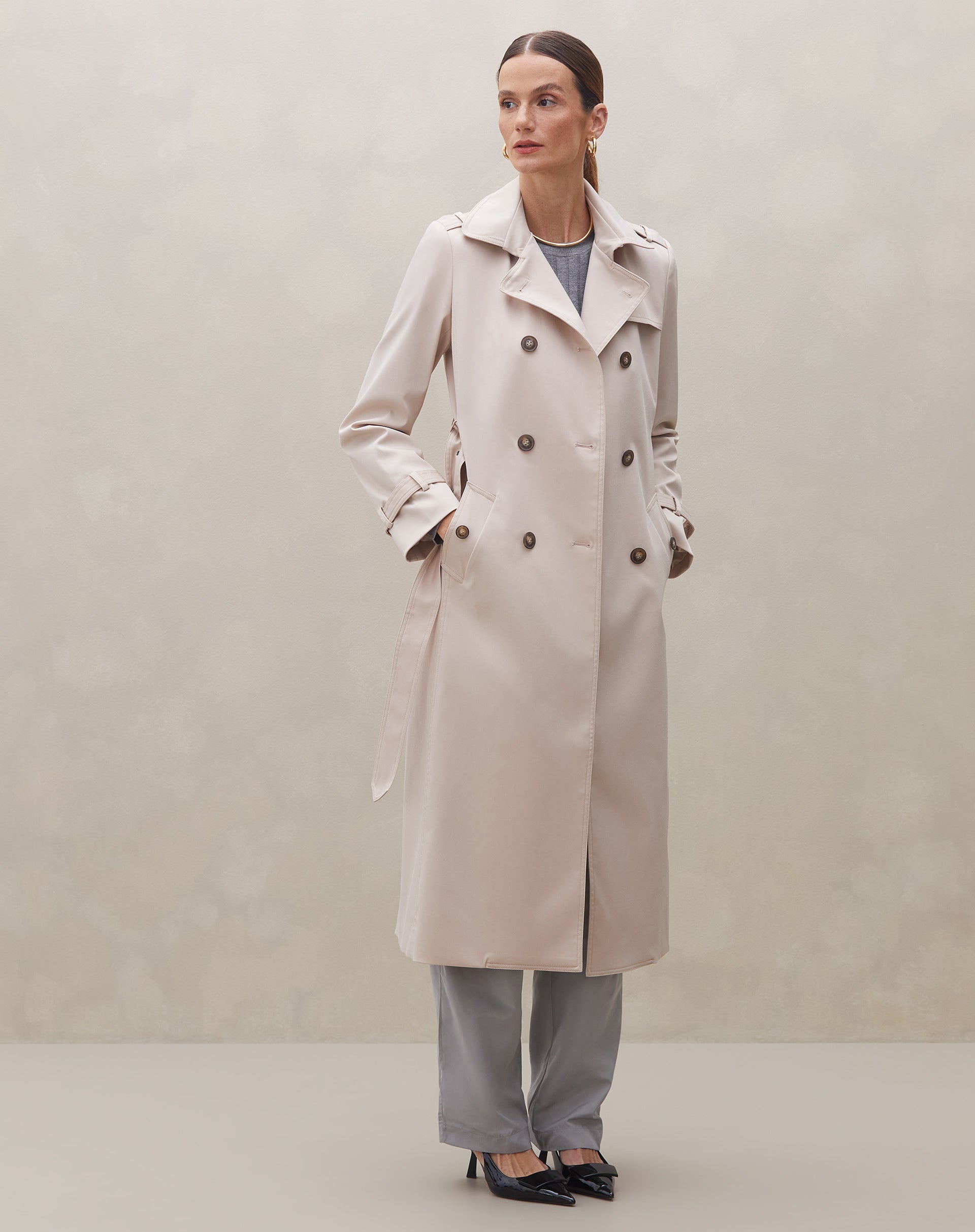 Modelo em pé vestindo trench coat bege com calças cinza e sapatos pretos.