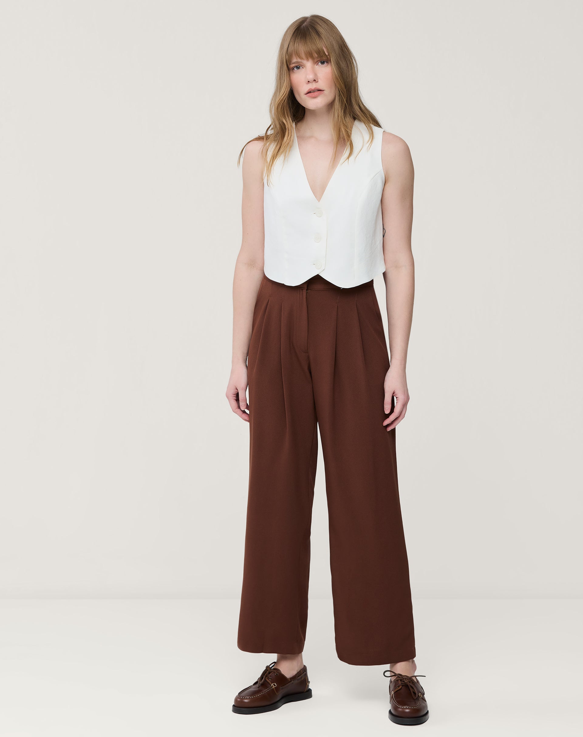Modelo em estúdio usando calça reta cropped marrom com pregas vista frontal, cós com meia-elástico e bolsos laterais