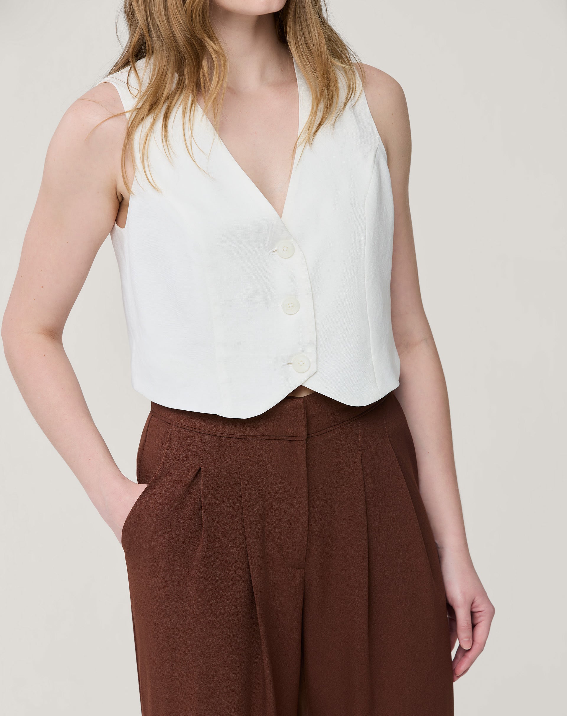 Modelo em pé com as mãos nos bolsos demonstrando o caimento e o comprimento cropped da calça marrom