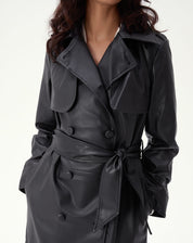 Vista frontal do trench coat preto PU com abotoamento duplo, gola clássica e cinto marcando a cintura, modelagem maxi em corpo inteiro.