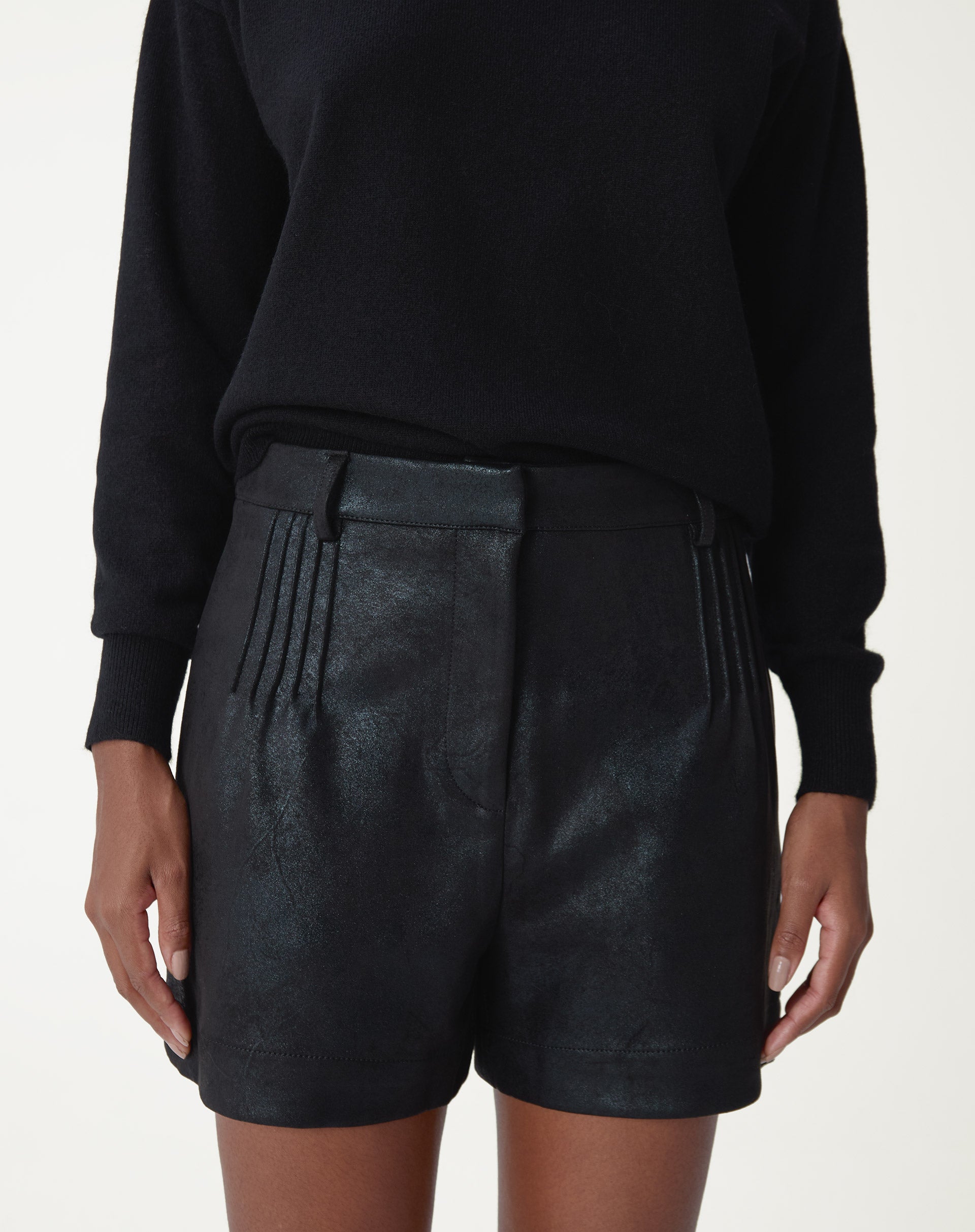 Modelo vista frontal usando shorts metalizado preto com pregas frontais e cós médio, combinado com suéter preto - detalhe de textura suedine