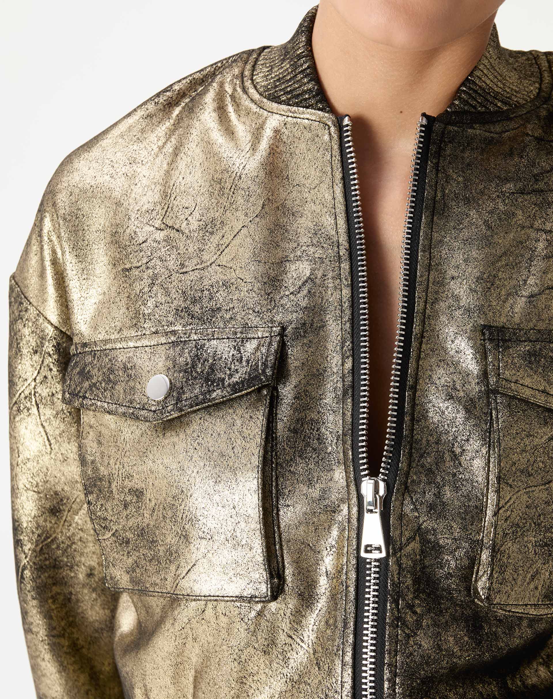 Close do peito da jaqueta bomber dourada mostrando bolso de aba com botão, textura metalizada e detalhe do zíper frontal