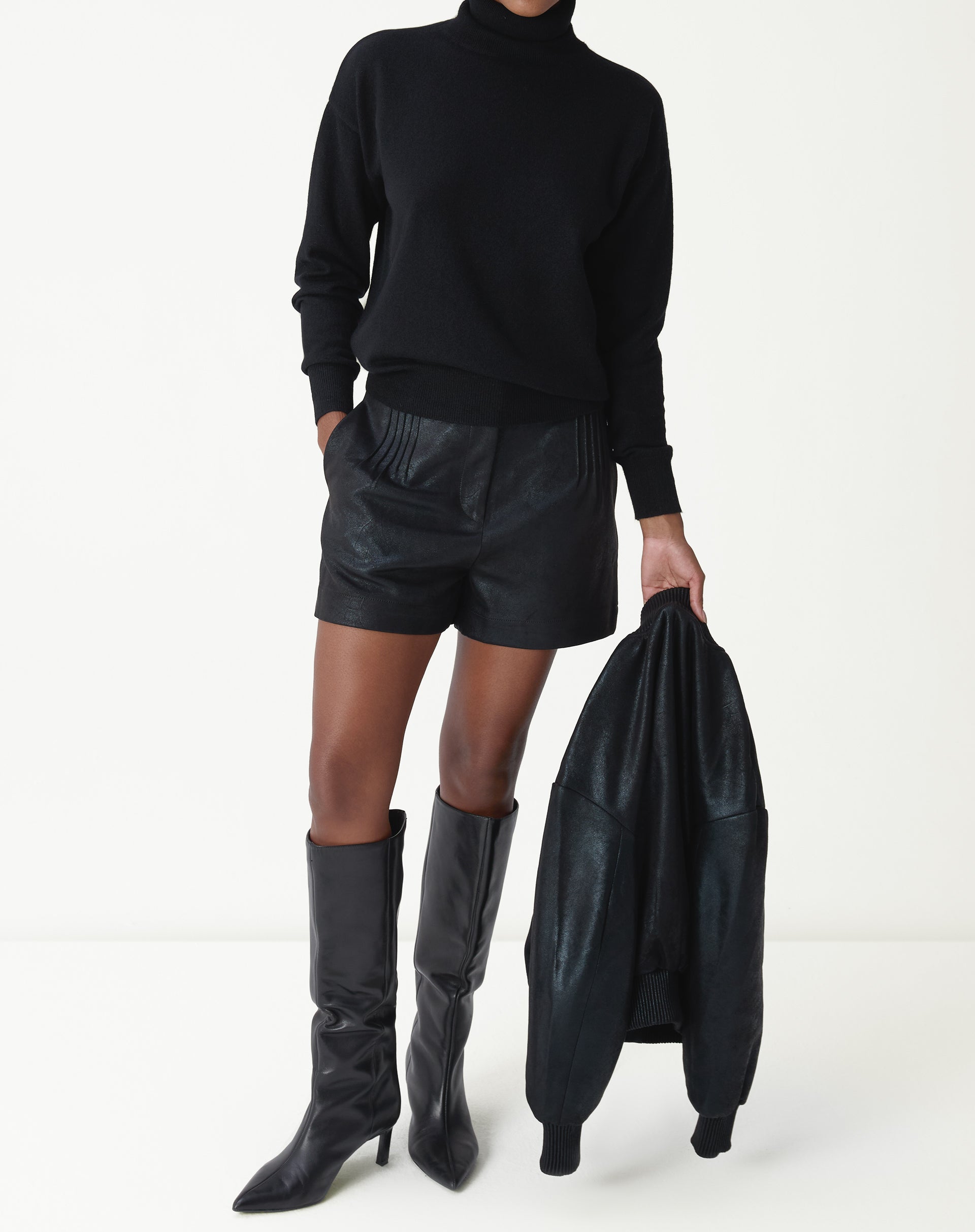 Mulher vestindo suéter gola alta de cashmere preto com shorts de couro e botas cano alto, look completo frontal