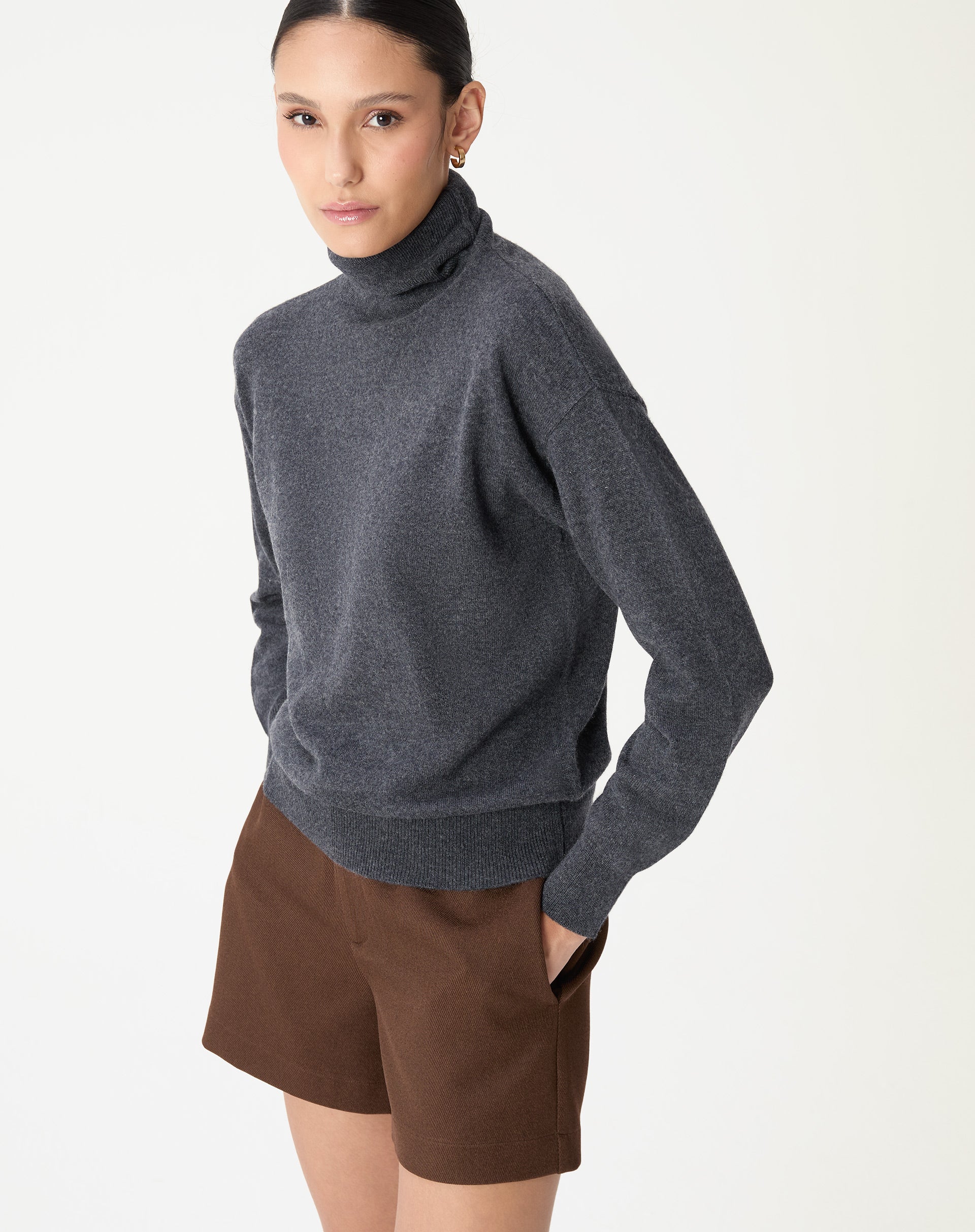 Modelo de lado vestindo um suéter de gola alta em cashmere cinza mescla escuro com um shorts marrom, caimento regular e estilo minimalista.