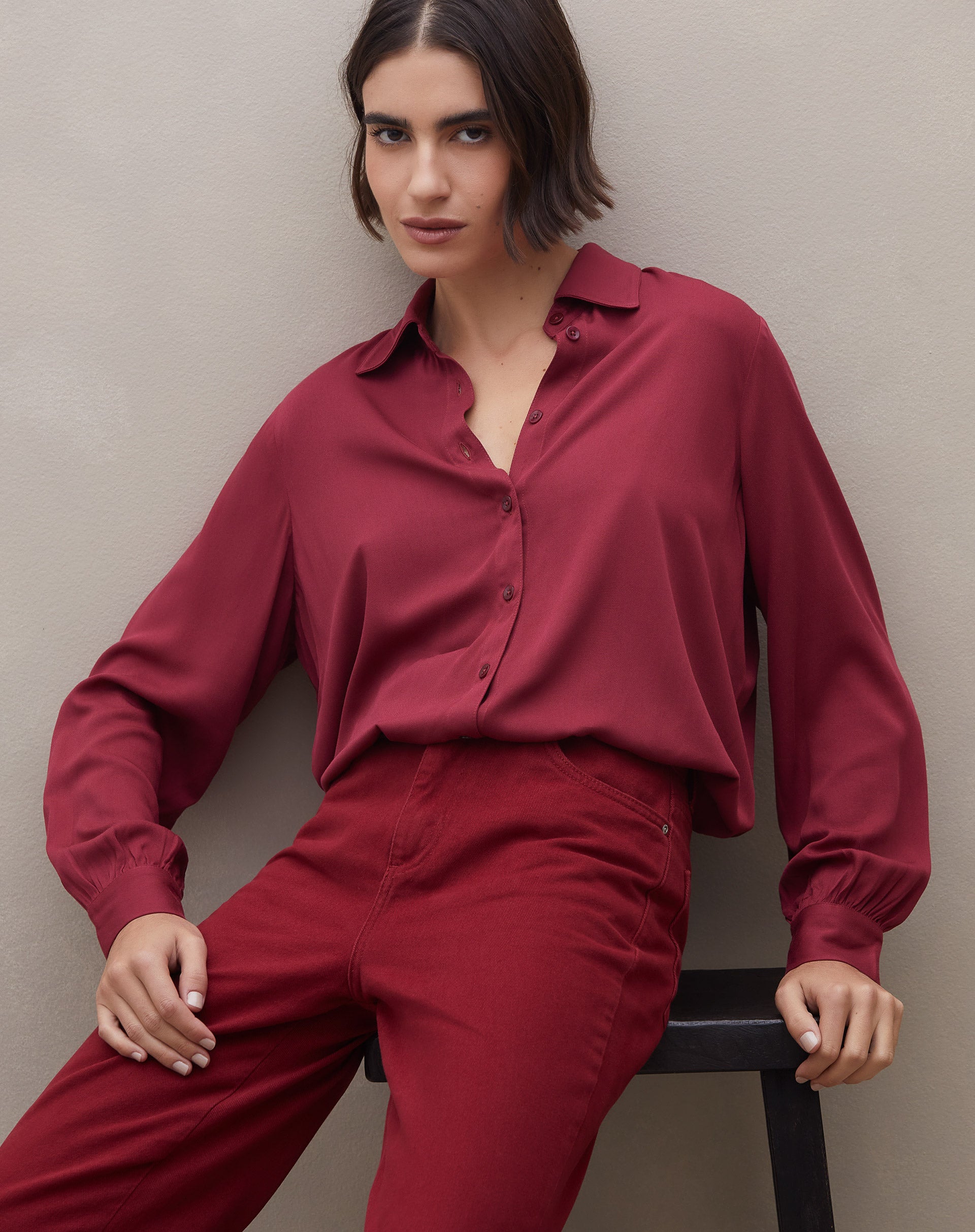Camisa feminina manga longa em viscose vinho, modelo sentada em look monocromático com calça vinho, mostrando caimento fluido